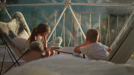 los niños miran fuera de la tienda transparente descansando las cabezas en las manos. niño y niña pensativos se relajan sentados en la mesa en el lugar de glamping. mañana en el complejo