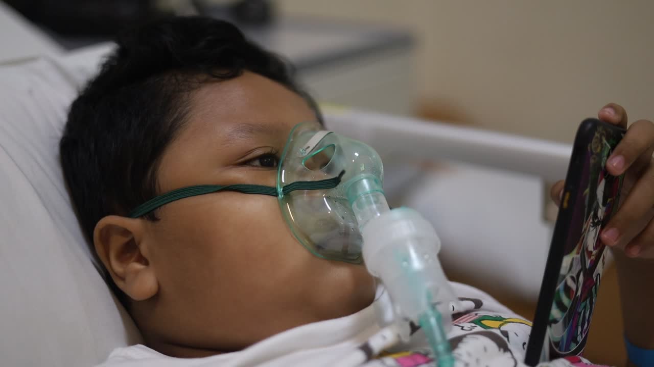 Primo piano di un adorabile bambino asiatico con maschera d'ossigeno sul viso a letto in ospedale mentre gioca con il cellulare