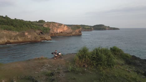 Aerial-flyover:-Male-motorcycle-tourist-enjoys-view-atop-ocean-cliff