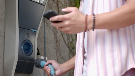 charging electric car, man holding charging cable and smartphone, focusing on task, in garage