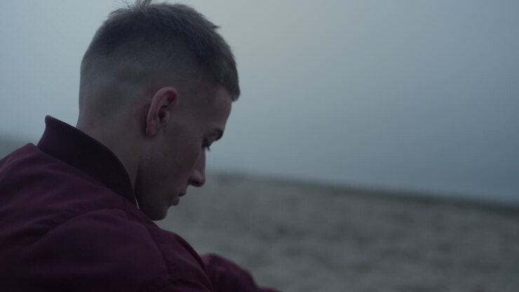 Sad man sitting sandy beach in morning. Young guy with upset face expression