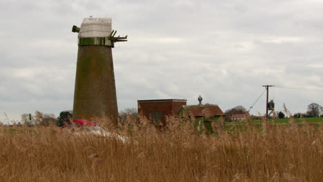 strzał boczny patrzący przez trzcinę na opuszczoną pompę wodną młyna wiatrowego norfolk broads z łodzią przechodzącą przez rzekę mrówkę w pobliżu mostu ludham