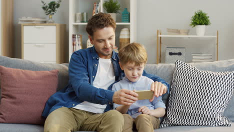 Vater-Und-Kleiner-Sohn-Verbringen-Zeit-Miteinander,-Mann-Bringt-Einem-Jungen-Bei,-Das-Smartphone-Zu-Hause-Im-Wohnzimmer-Zu-Benutzen