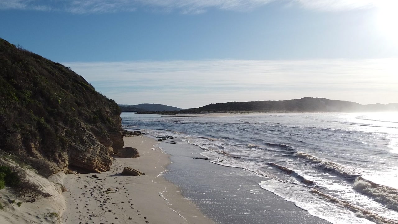 Gentle waves lap against a sandy beach backed by rugged cliffs under a clear blue sky.