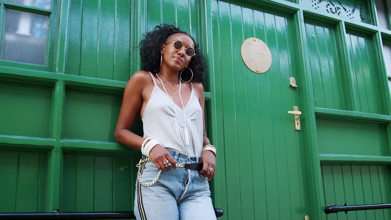 Fashionable young black woman wearing sunglasses leaning by green door outdoors lifts sunglasses, low angle