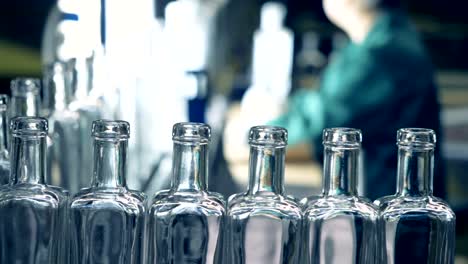 a straight line of glassy bottles is moving along the conveyor while a factory worker is observing the process