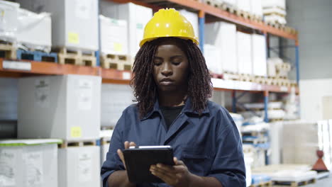 focused african american female employee working in warehouse, counting goods using tablet