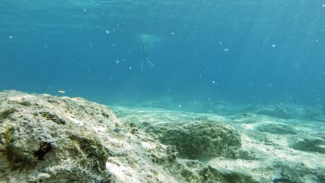 Ein-Mann,-Der-Auf-Dem-Kristallklaren-Wasser-Des-Strandes-Von-Paralia-Emplisi-Freitaucht,-Während-Die-Sonnenstrahlen-Hindurchstrahlen-Und-Einige-Fische-Herumschwimmen