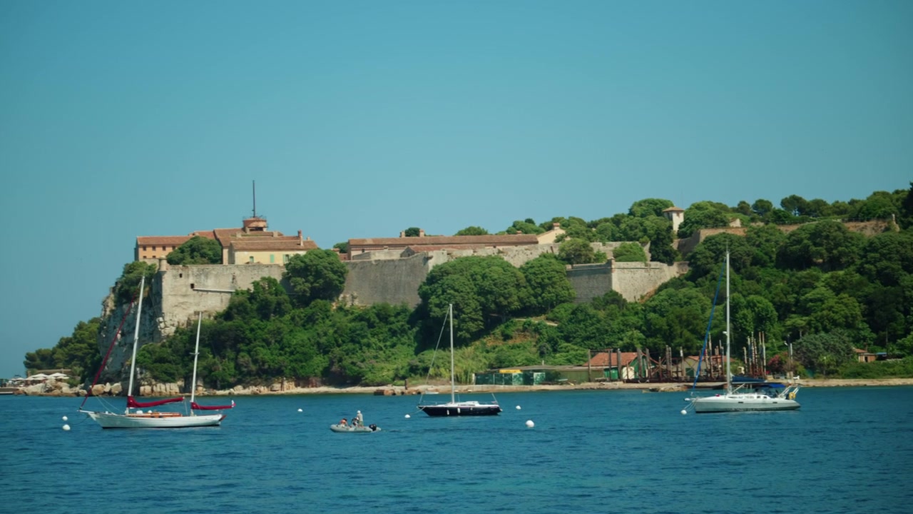Catamarano bianco che naviga in acque tranquille e di un blu profondo con una costa verde lussureggiante e case tradizionali sullo sfondo sotto un cielo sereno.