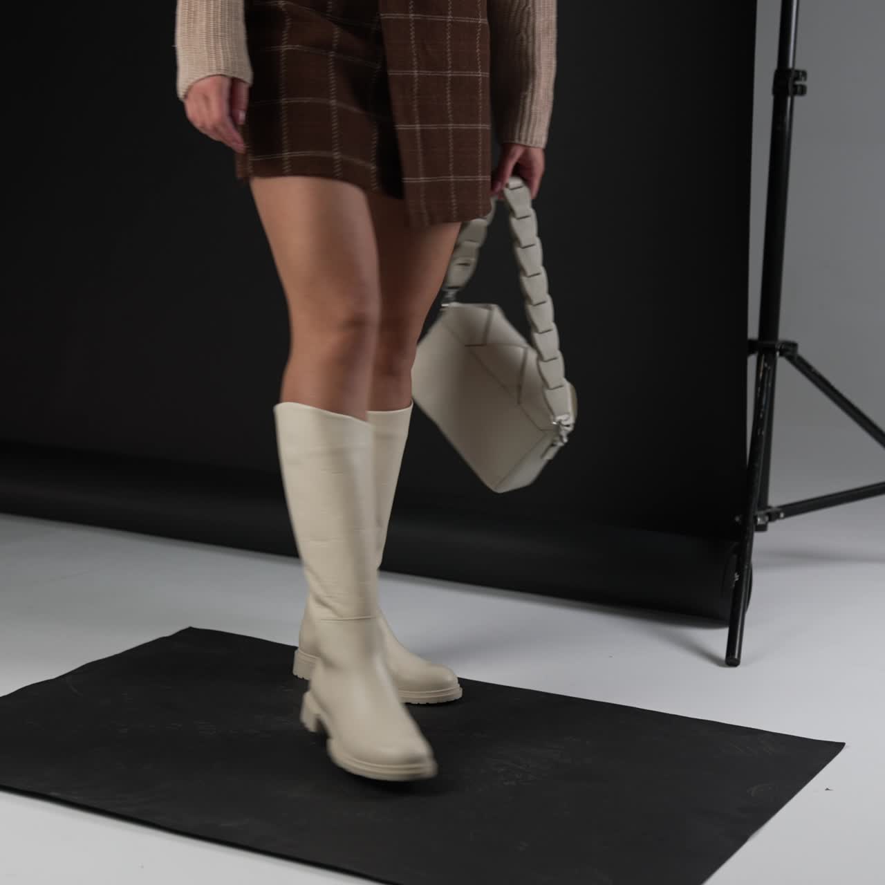 Female demonstrating milk-white high boots on plain soles and same-shade bag. Model turns to show the footwear from different sides. Studio backdrop