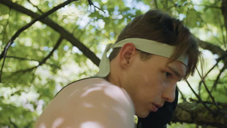 side profile determined tourist in headband throwing relentless punches at tree bark while scanning ahead in leafy forest, sweat on skin, muscles tensed in focused training drill under sunlight