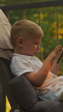 little boy plays on contemporary smartphone enjoying pleasant evening. amazing nature scenery surrounds toddler child sitting in comfortable armchair at sunset