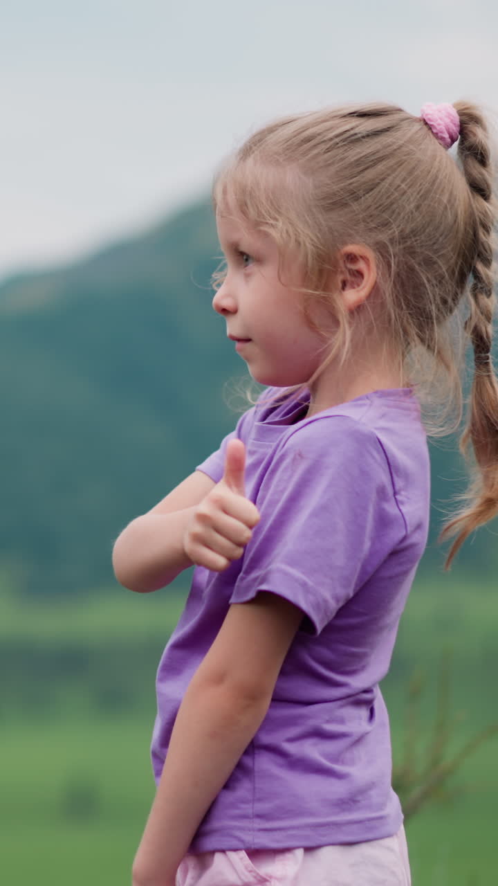 adorable niña con trenza muestra el pulgar hacia arriba de pie sobre el valle verde contra grandes montañas distantes en el día nublado vista lateral en cámara lenta