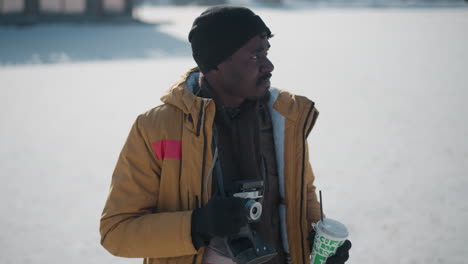 lensman sipping morning coffee under winter sky, holding camera in one hand while scanning urban landscape shining bright in sunlight, visible breath in crisp cold air, bag hanging at chest