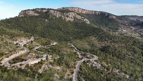 aerial approaching shot of mountain range in esporles with serpentine road on hill during sunny day, mallorca