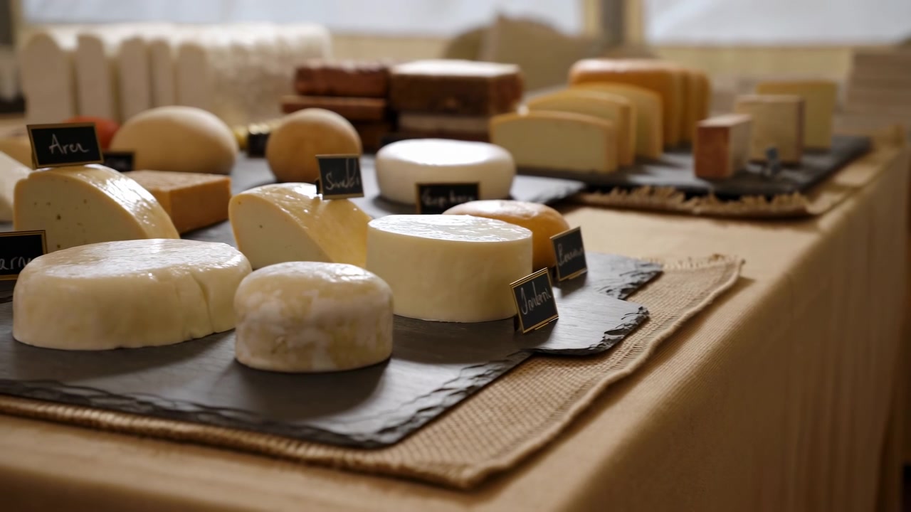 Une vue rapprochée en mouvement lent révèle une présentation de fromages artisanaux disposés sur des planches d'ardoise foncée, sur un chemin de table en toile de jute et une nappe beige, avec des meules entières, des demi-meules, des quartiers, et une lumière de marché chaleureuse.