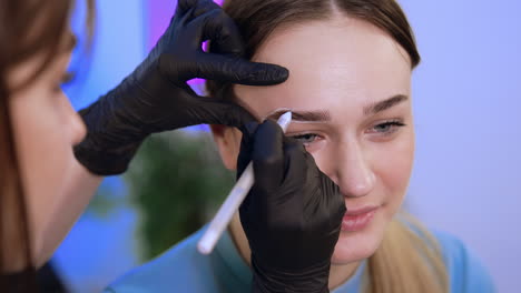 young pretty lady is prepared for eyebrow permanent. female beauty specialist draws the contour with white pencil. close up.