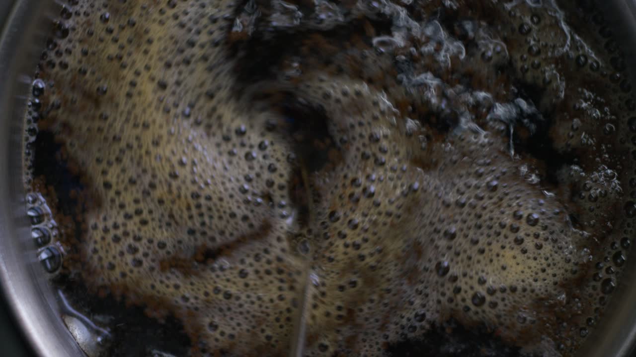 A top view close-up of tea boiling with a foamy surface and bubbles, and someone stirring it with a spoon