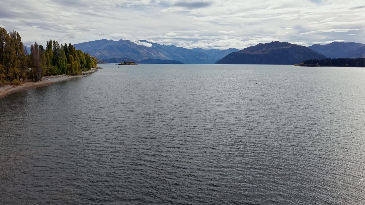 Aerial drone footage flying over Lake Wanaka in New Zealand, showing calm water, green trees, distant mountains and cloudy sky