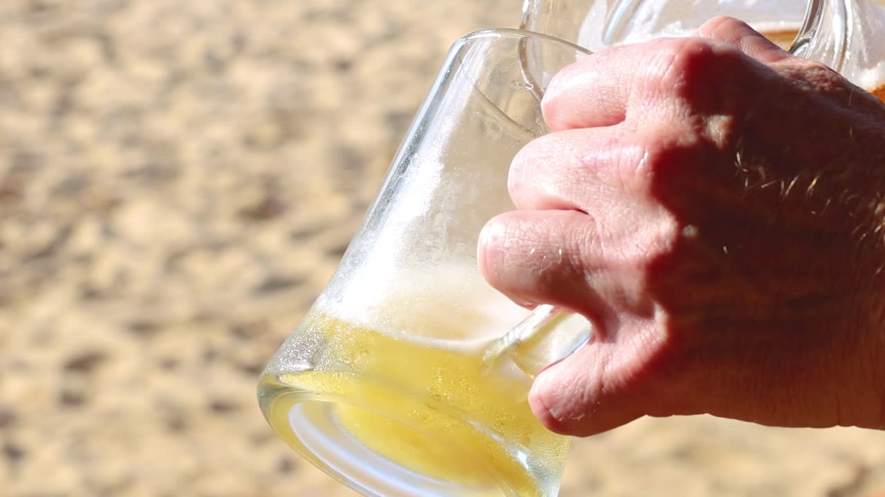 A hand holding a glass jug, pouring frothy beer with a sandy background under bright sunlight.