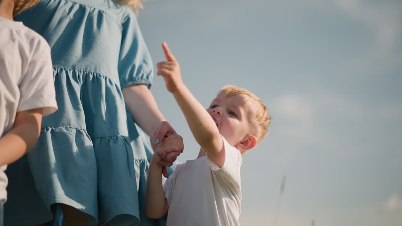 A little boy in a white shirt smiles pointing at the environment, holding his mother s hand, who is wearing a blue dress. Another boy is partially visible in the background