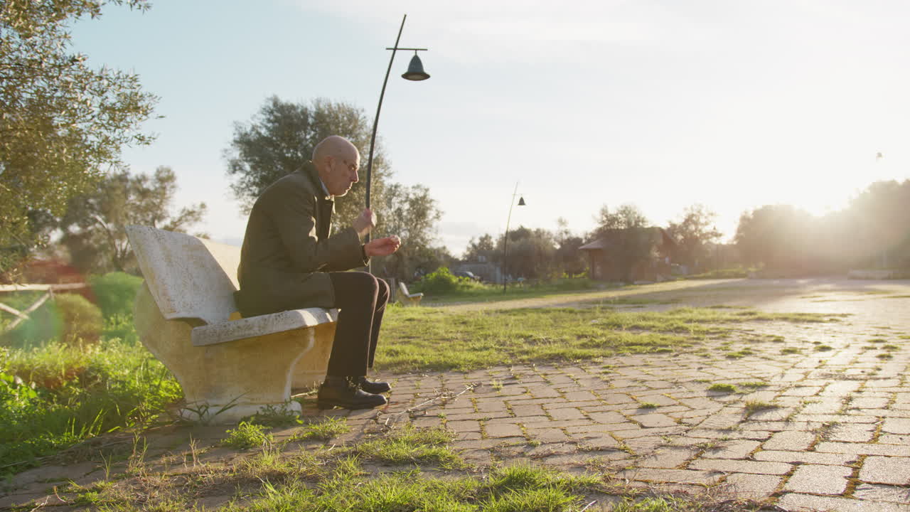 Old Man Relaxes on a Park Bench Under the Sun Eating a Snack