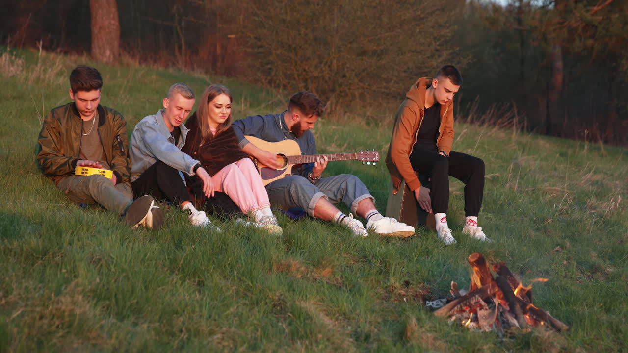 Young people sitting on the hill in front of campfire. Friends having good time with singing along with playing guitar and other instruments.
