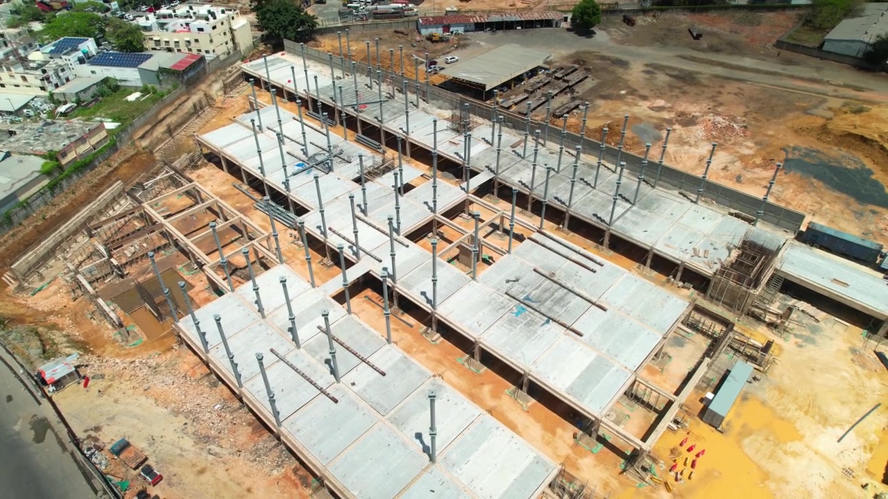Rekaman drone udara sudut tinggi terbang maju dan mengorbit ke kanan di atas lokasi konstruksi gedung komersial besar, menunjukkan pilar baja, lempengan lantai beton, dan sekitarnya.