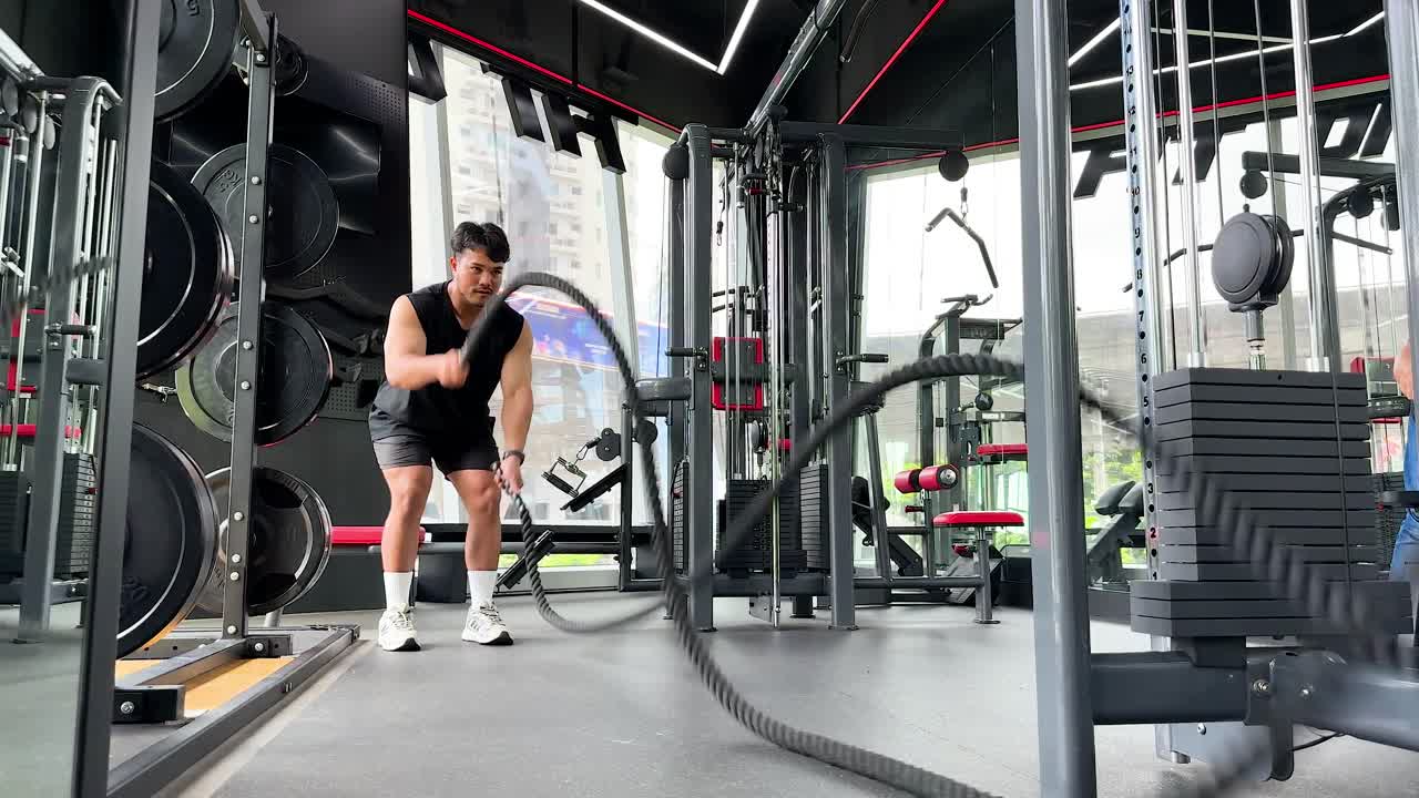 A person performs a vigorous battle rope exercise in a well-equipped gym with bright lighting and dynamic camera angles