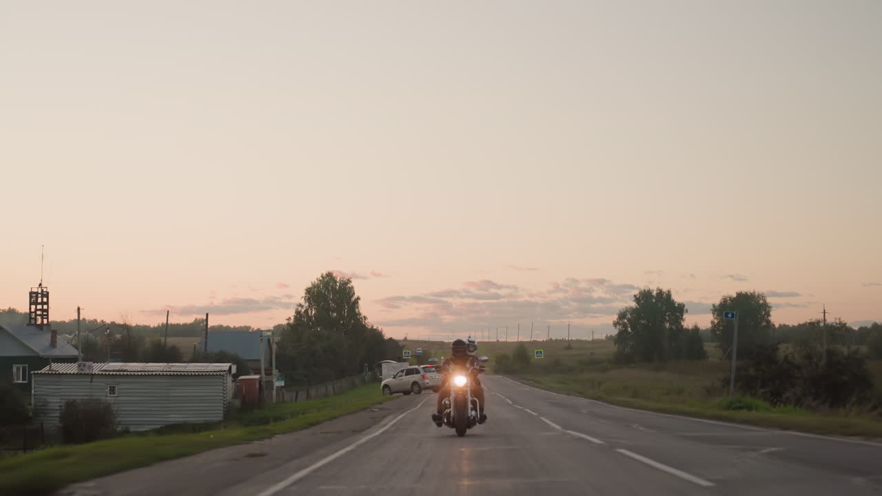 Motorcyclist wearing helmet rides on open countryside road behind jeep turning ahead during scenic sunset, with glowing headlight, peaceful horizon, and rural landscape
