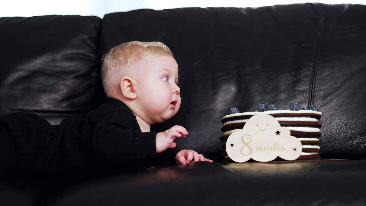 Lovely Caucasian baby boy with plump cheeks lies on the black leather sofa. Cute child is focused on a cake in front of him.
