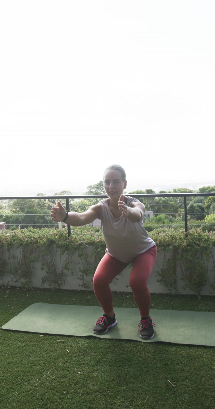 Video verticale: Donna adulta che fa squat da una posizione profonda su un tappetino sul balcone, in leggings rossi, per allenarsi.