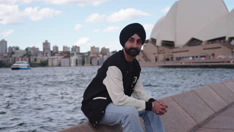 indian man in turban sitting in front of famous sydney opera house in sydney, australia