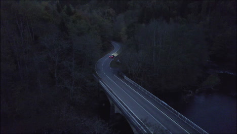 Night-and-dark-drone-shot-of-a-car-passing-by-with-headlights-on-a-swirly-road-in-between-the-woods-and-driving-away-LOG
