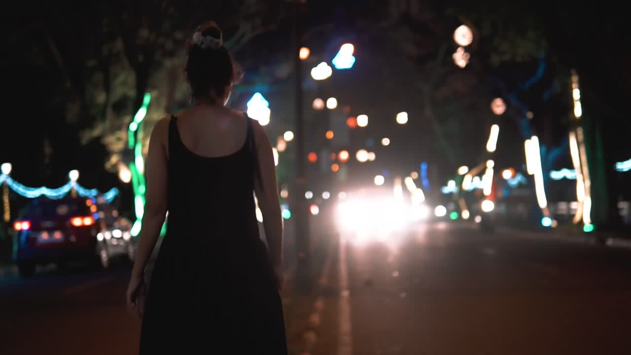 chica con un vestido negro caminando al lado de la carretera con tráfico entrante durante la noche