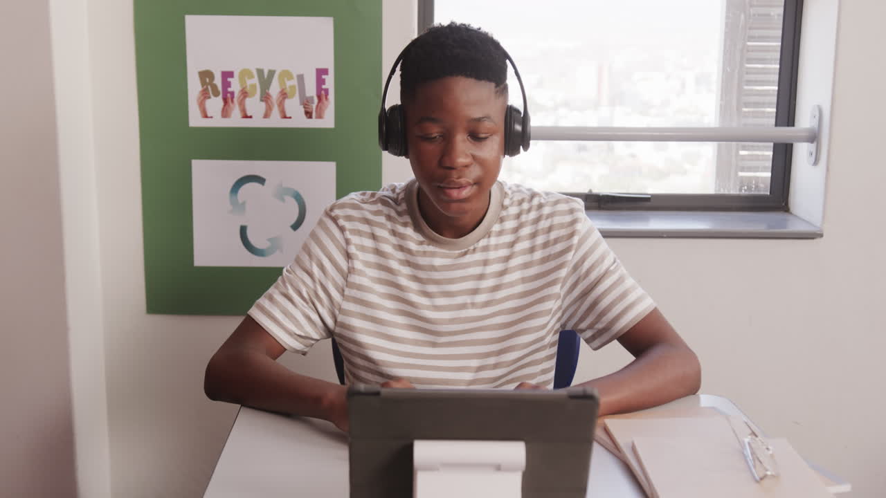 In school, African American teenage boy wearing headphones using tablet for studying at desk