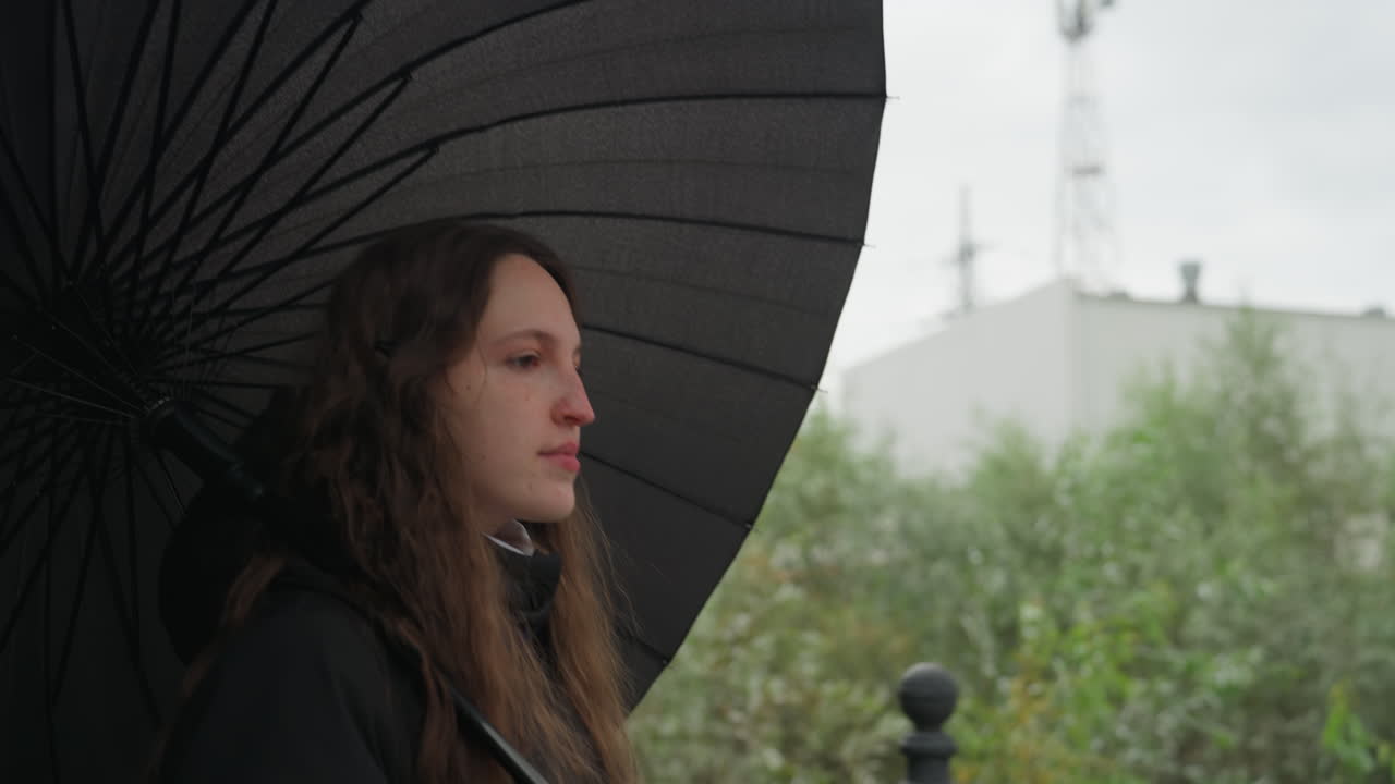 Donna con ombrello che cammina in una strada urbana nuvolosa, ritratto di profilo che rivela uno stato d'animo contemplativo, lunghi capelli ondulati, cappotto scuro, silhouette di lampione, edificio industriale distante, fogliame verde, delicato