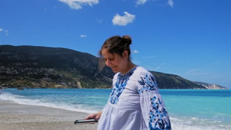 Mujer-Encantadora-Con-Teléfono-Inteligente-En-La-Mano-Alejándose-De-La-Orilla-Sonriendo-En-La-Playa-De-Agia-Kiriaki,-Zola-En-Cefalonia,-Grecia