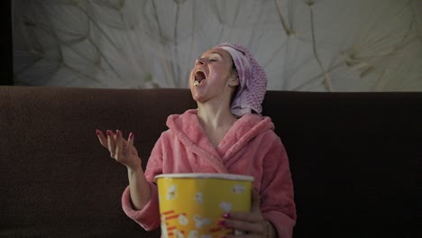 woman watching a late night movie at tv, eating popcorn. bathrobe, facial mask