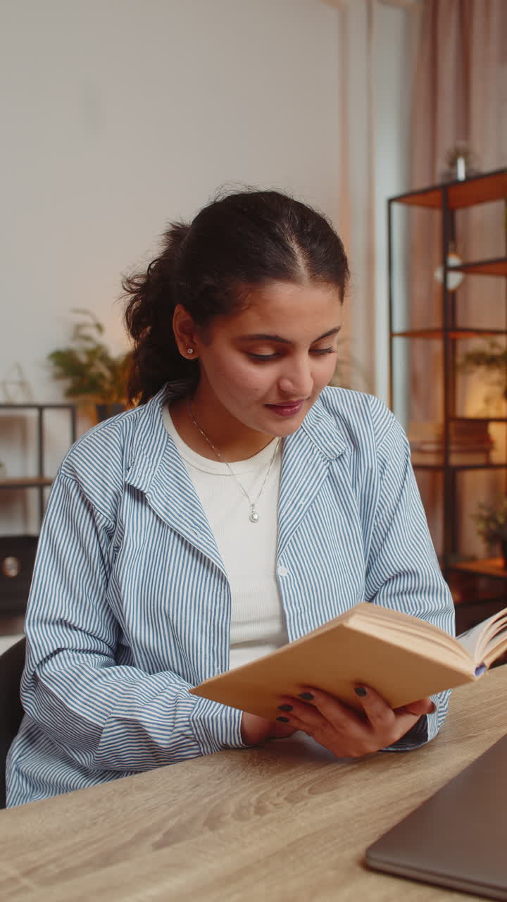Indian young woman reading book turning pages smiling enjoying literature taking rest sits at table