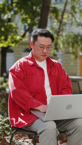 Asian-man-freelancer-working-online-distant-job-with-laptop-browsing-website-sitting-on-city-street