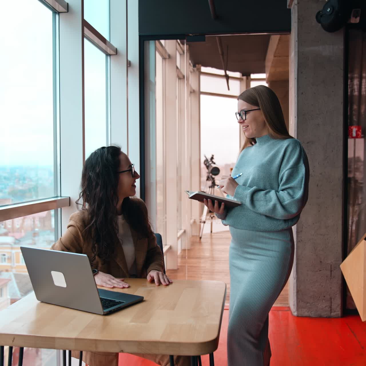 Two positive female workers communicating in the office. Blonde pregnant lady holding a notebook and taking notes. Brunette colleague sits in front of laptop