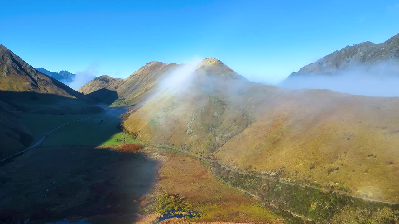 Drone footage captures fog rolling over sunlit mountains near Moke Lake, Queenstown. Clear skies and vibrant colors create a serene atmosphere