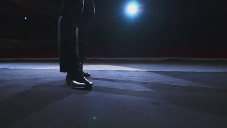 actor's legs dancing on stage. man in black shoes dancing against the theatre hall at dark scenic light. close-up.