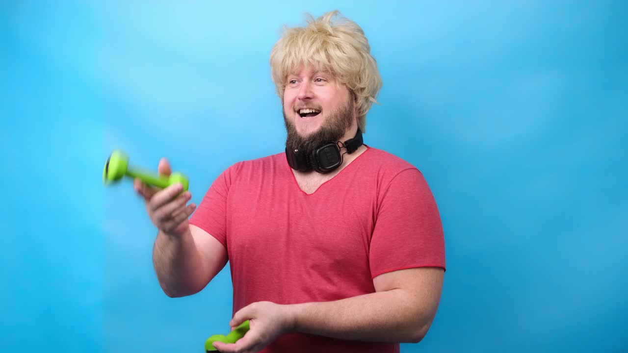 웃긴 남자는 헤드폰을 입고 70의 죄에서 머리카락을 고 작은 녹색 덤벨과 미소를 가진 분홍색 티셔츠를 다루고 있습니다.