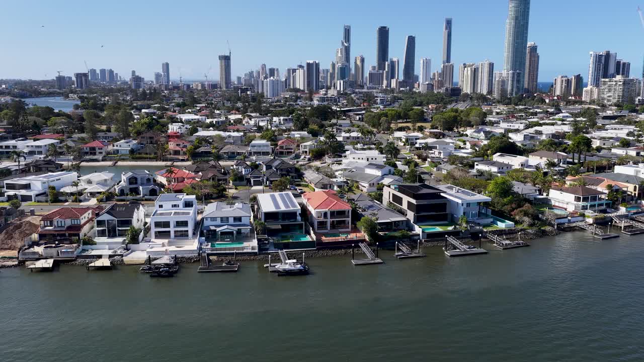 Drone glides above riverfront houses toward city skyscrapers under bright daylight, steady wide perspective