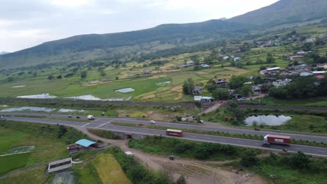 cinematic aerial drone shot circling around passing cars and heavy transportation vehicles on the four lanes indian highway and railway track beside it surrounded by small village and mountains