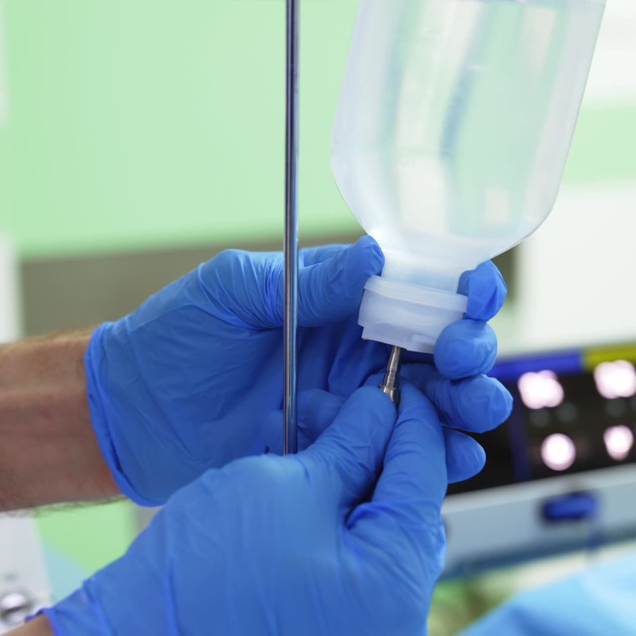 Hands in blue gloves attaching medicines bottle to the injection drip. Medical equipment in blur at the background