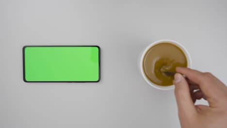 man hand cup of hot coffee and using smartphone watching green screen top view. smartphone with green mock-up screen business concept. person hand stirring coffee with spoon on table. slow motion.