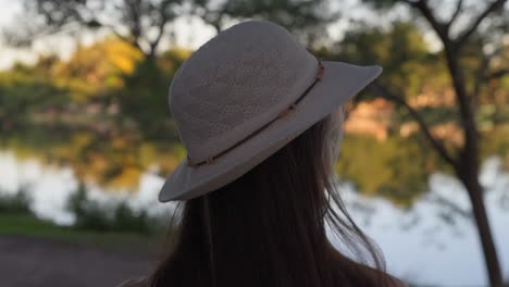 beautiful young woman walking carefree near lake while putting on white hat in nature at sunset, close up